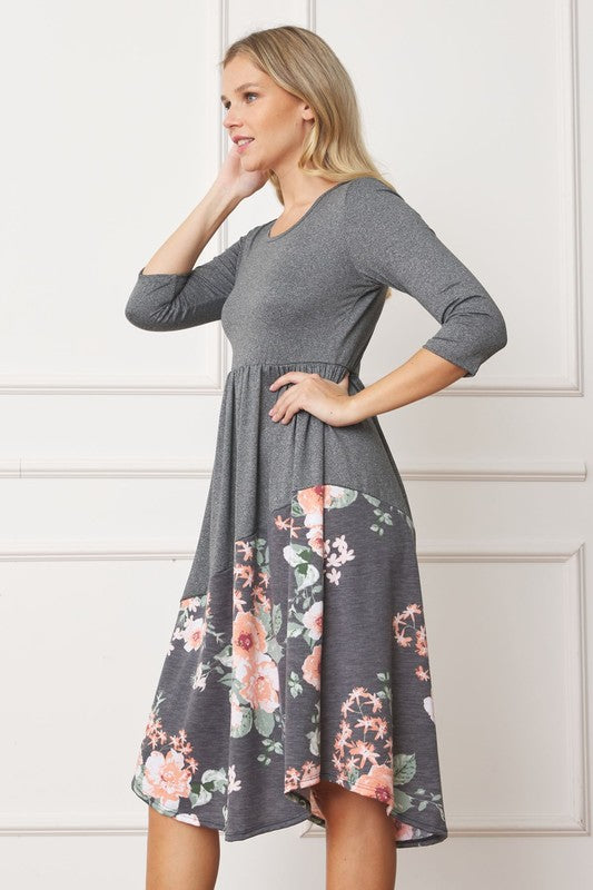 A woman stands in front of a white wall wearing the Asymmetrical Accent Midi Dress, which features a colorful floral patterned skirt. She smiles while resting her left hand on her hip and keeping her right arm by her side. Her outfit, crafted from polyester and spandex, is elegantly finished with black boots.