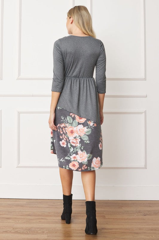 A woman stands in front of a white wall wearing the Asymmetrical Accent Midi Dress, which features a colorful floral patterned skirt. She smiles while resting her left hand on her hip and keeping her right arm by her side. Her outfit, crafted from polyester and spandex, is elegantly finished with black boots.