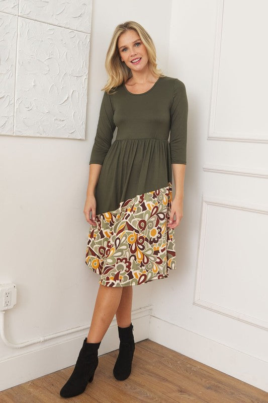 A woman stands in front of a white wall wearing the Asymmetrical Accent Midi Dress, which features a colorful floral patterned skirt. She smiles while resting her left hand on her hip and keeping her right arm by her side. Her outfit, crafted from polyester and spandex, is elegantly finished with black boots.