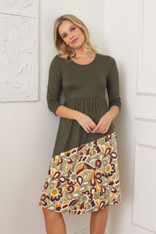A woman stands in front of a white wall wearing the Asymmetrical Accent Midi Dress, which features a colorful floral patterned skirt. She smiles while resting her left hand on her hip and keeping her right arm by her side. Her outfit, crafted from polyester and spandex, is elegantly finished with black boots.