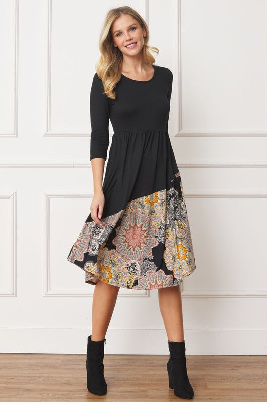 A woman stands in front of a white wall wearing the Asymmetrical Accent Midi Dress, which features a colorful floral patterned skirt. She smiles while resting her left hand on her hip and keeping her right arm by her side. Her outfit, crafted from polyester and spandex, is elegantly finished with black boots.