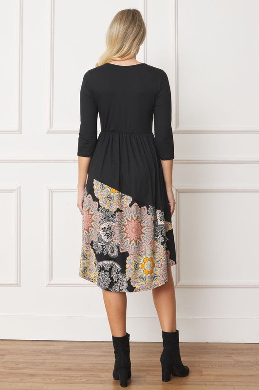 A woman stands in front of a white wall wearing the Asymmetrical Accent Midi Dress, which features a colorful floral patterned skirt. She smiles while resting her left hand on her hip and keeping her right arm by her side. Her outfit, crafted from polyester and spandex, is elegantly finished with black boots.