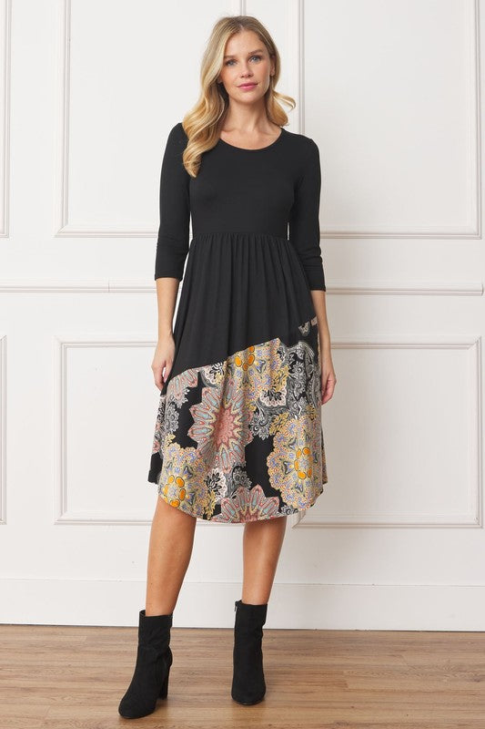 A woman stands in front of a white wall wearing the Asymmetrical Accent Midi Dress, which features a colorful floral patterned skirt. She smiles while resting her left hand on her hip and keeping her right arm by her side. Her outfit, crafted from polyester and spandex, is elegantly finished with black boots.