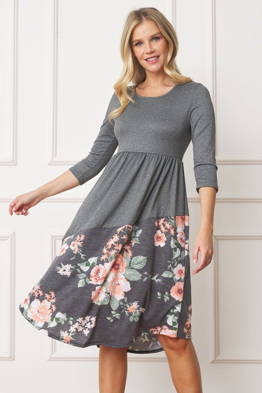 A woman stands in front of a white wall wearing the Asymmetrical Accent Midi Dress, which features a colorful floral patterned skirt. She smiles while resting her left hand on her hip and keeping her right arm by her side. Her outfit, crafted from polyester and spandex, is elegantly finished with black boots.