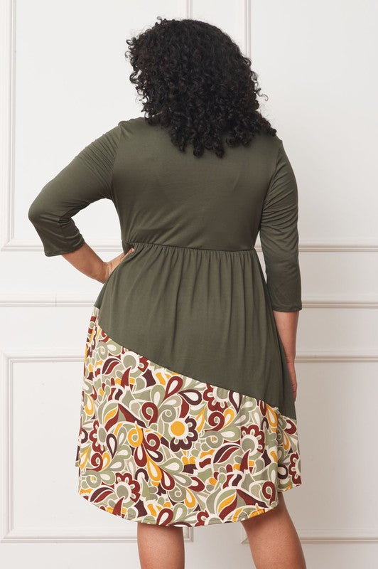 A person standing in front of a white wall, wearing a Plus Asymmetrical Accent Midi Dress made of polyester spandex with three-quarter sleeves and a skirt featuring a colorful floral pattern.