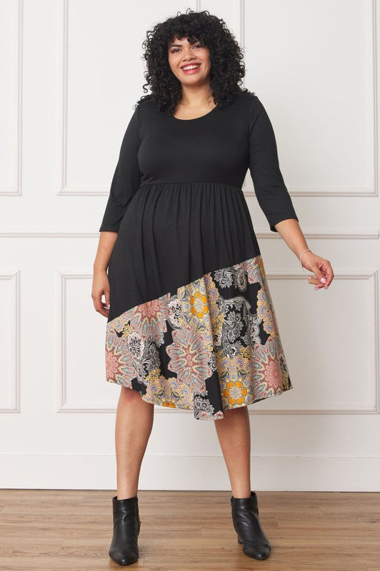 A person standing in front of a white wall, wearing a Plus Asymmetrical Accent Midi Dress made of polyester spandex with three-quarter sleeves and a skirt featuring a colorful floral pattern.