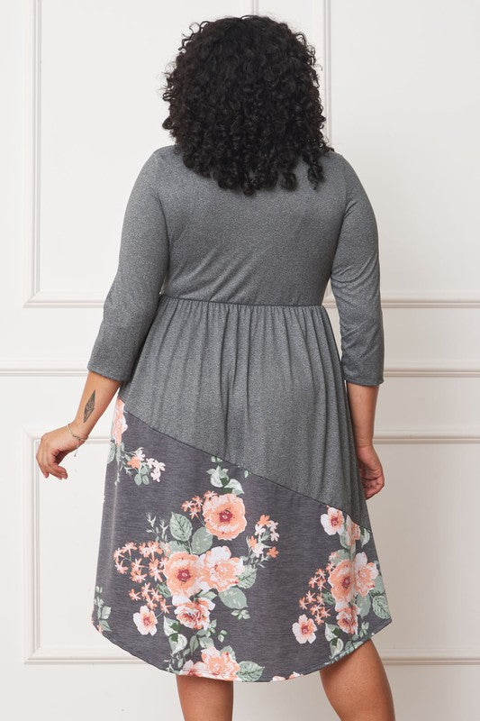 A person standing in front of a white wall, wearing a Plus Asymmetrical Accent Midi Dress made of polyester spandex with three-quarter sleeves and a skirt featuring a colorful floral pattern.
