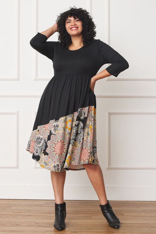 A person standing in front of a white wall, wearing a Plus Asymmetrical Accent Midi Dress made of polyester spandex with three-quarter sleeves and a skirt featuring a colorful floral pattern.