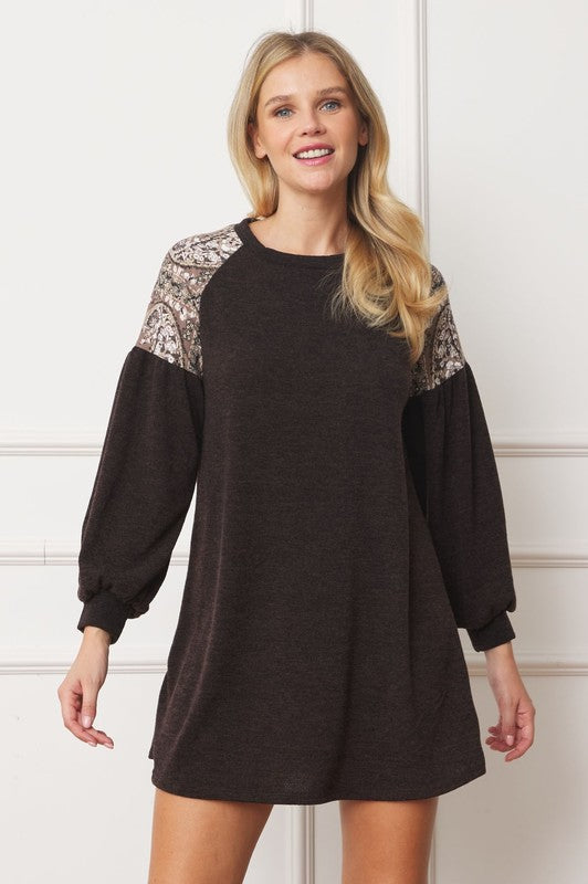 A person with long blonde hair smiles while wearing a dark Paisley Shoulder Accent Tunic Dress, featuring delicate lace detail on the shoulders. Made in the United States, this dress offers added comfort with its blend of 95% polyester and 5% spandex.