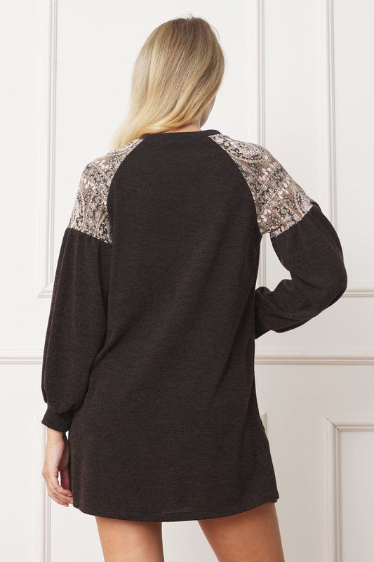 A woman with shoulder-length brown hair wears the "Plus Paisley Shoulder Accent Tunic Dress," which is loose-fitting and black with paisley shoulder accents. She stands in front of a plain, light-colored background.