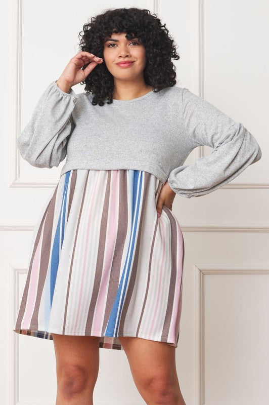 A person wearing a black hat, a dark gray Balloon Sleeve Pullover Dress, a multicolored striped skirt, and black knee-high boots poses in front of a white paneled wall. The entire ensemble exudes elegance and style, reflecting the craftsmanship made in the United States.