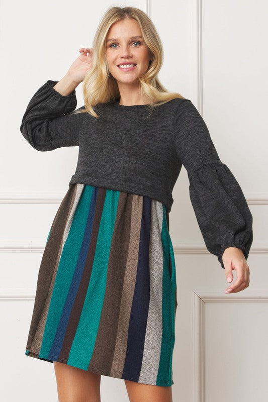 A woman with curly hair, dressed in a Plus Balloon Sleeve Pullover Dress featuring a gray top and vertical stripes in various colors on the high-waisted skirt, stands against a plain background. Her attire includes elements of Polyester Spandex for comfort and style.