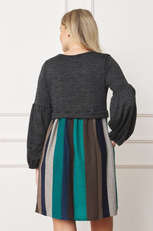 A person wearing a black hat, a dark gray Balloon Sleeve Pullover Dress, a multicolored striped skirt, and black knee-high boots poses in front of a white paneled wall. The entire ensemble exudes elegance and style, reflecting the craftsmanship made in the United States.