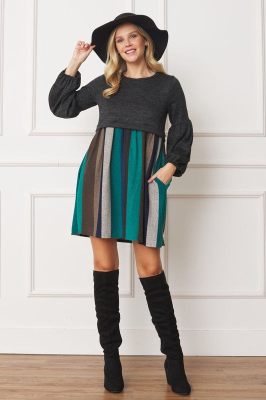 A person wearing a black hat, a dark gray Balloon Sleeve Pullover Dress, a multicolored striped skirt, and black knee-high boots poses in front of a white paneled wall. The entire ensemble exudes elegance and style, reflecting the craftsmanship made in the United States.