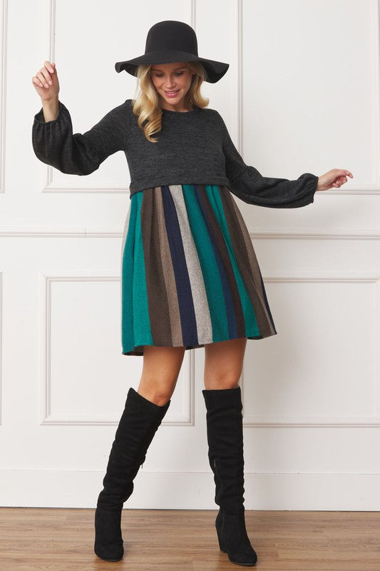 A person wearing a black hat, a dark gray Balloon Sleeve Pullover Dress, a multicolored striped skirt, and black knee-high boots poses in front of a white paneled wall. The entire ensemble exudes elegance and style, reflecting the craftsmanship made in the United States.