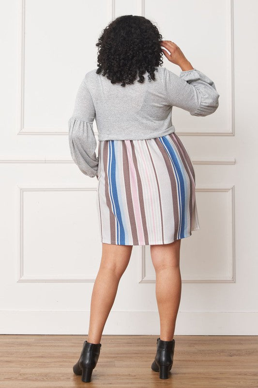 A woman with curly hair, dressed in a Plus Balloon Sleeve Pullover Dress featuring a gray top and vertical stripes in various colors on the high-waisted skirt, stands against a plain background. Her attire includes elements of Polyester Spandex for comfort and style.