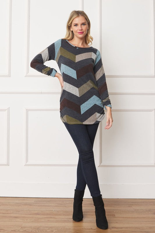 A woman with shoulder-length blonde hair is wearing a Shirred Side Accent Top that features chevron stripes in blue, gray, and olive. The long-sleeve top, made from a polyester-spandex blend, complements her figure perfectly as she stands in front of a white background.