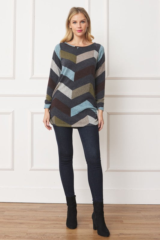 A woman with blonde hair is wearing a Shirred Side Accent Top, showcasing a multicolored chevron pattern, paired with dark jeans while standing against a white paneled backdrop. The top, made from 95% polyester and 5% spandex, elegantly complements her look.