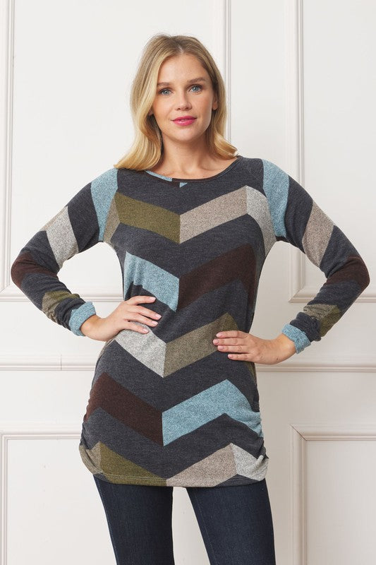 A woman with shoulder-length blonde hair is wearing a Shirred Side Accent Top that features chevron stripes in blue, gray, and olive. The long-sleeve top, made from a polyester-spandex blend, complements her figure perfectly as she stands in front of a white background.