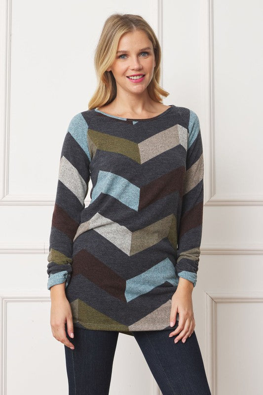 A woman with shoulder-length blonde hair is wearing a Shirred Side Accent Top that features chevron stripes in blue, gray, and olive. The long-sleeve top, made from a polyester-spandex blend, complements her figure perfectly as she stands in front of a white background.