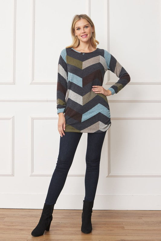 A woman wearing a Shirred Side Accent Top, which is a long-sleeve shirt adorned with a chevron pattern in shades of blue, green, brown, and gray, stands against a white paneled background. The blend of polyester and spandex fabric ensures both comfort and style.