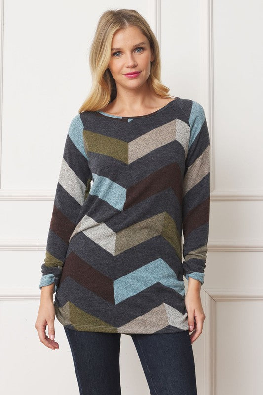 A woman with blonde hair is wearing a Shirred Side Accent Top, showcasing a multicolored chevron pattern, paired with dark jeans while standing against a white paneled backdrop. The top, made from 95% polyester and 5% spandex, elegantly complements her look.