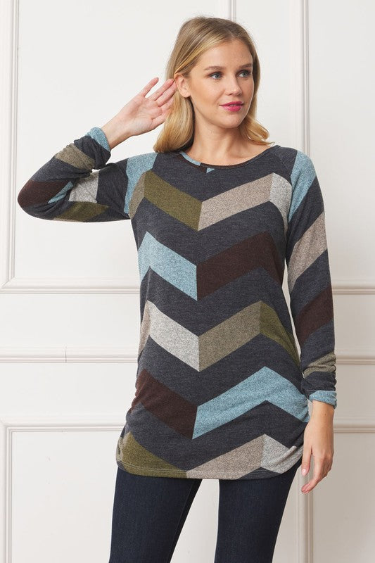 A woman with blonde hair is wearing a Shirred Side Accent Top, showcasing a multicolored chevron pattern, paired with dark jeans while standing against a white paneled backdrop. The top, made from 95% polyester and 5% spandex, elegantly complements her look.