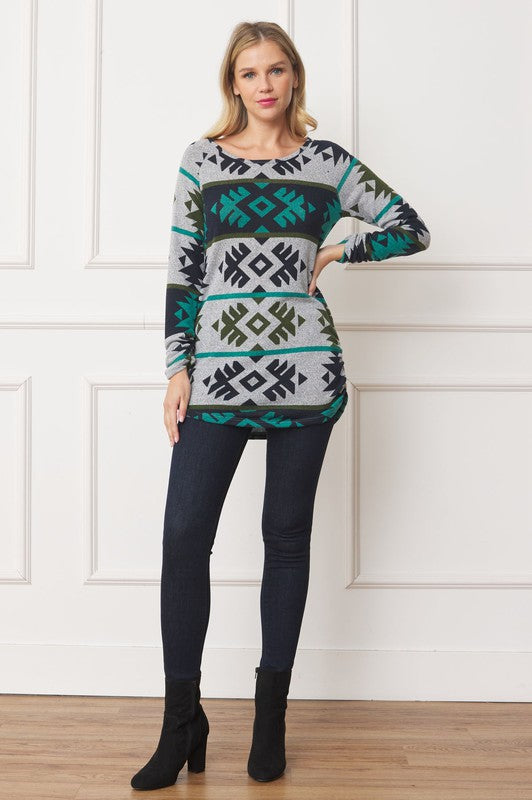 A woman stands against a white paneled wall, wearing an Aztec Shirred Side Accent Top. The top is a long-sleeve grey blend of polyester and spandex featuring green, black, and turquoise geometric patterns. She is looking to her right with her left hand near her face.
