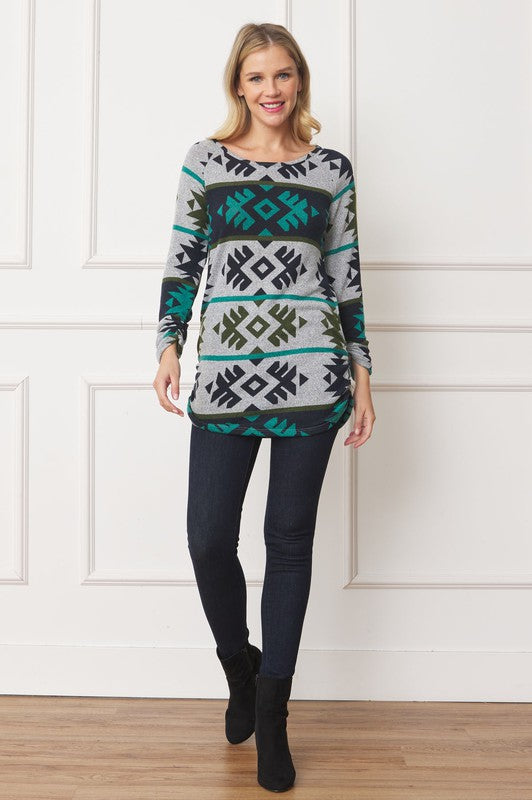 A woman stands against a white background, smiling. She wears the Aztec Shirred Side Accent Top made in the United States, featuring geometric designs in green, black, and white. The top is stylishly paired with dark jeans, and her right hand rests on her hip.