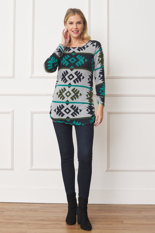 A woman stands against a white background, smiling. She wears the Aztec Shirred Side Accent Top made in the United States, featuring geometric designs in green, black, and white. The top is stylishly paired with dark jeans, and her right hand rests on her hip.
