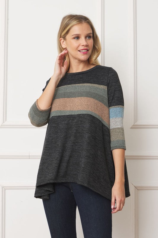 A woman with shoulder-length hair is wearing the Knit Stripe Panel A-Line Tunic, featuring horizontal brown, green, and blue knit stripe panels. She stands against a paneled white wall with one hand touching her neck and pairs the tunic with dark jeans.