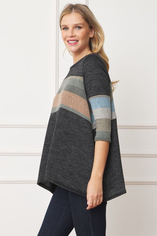 A woman with shoulder-length hair is wearing the Knit Stripe Panel A-Line Tunic, featuring horizontal brown, green, and blue knit stripe panels. She stands against a paneled white wall with one hand touching her neck and pairs the tunic with dark jeans.