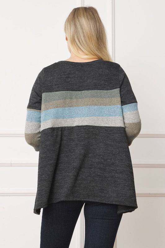 A person is standing against a beige backdrop, wearing a Plus Knit Stripe Panel A Line Tunic with horizontal stripes in various shades and blue jeans.