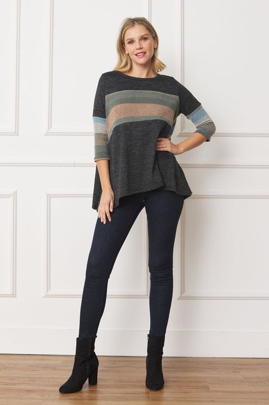 A woman with shoulder-length hair is wearing the Knit Stripe Panel A-Line Tunic, featuring horizontal brown, green, and blue knit stripe panels. She stands against a paneled white wall with one hand touching her neck and pairs the tunic with dark jeans.