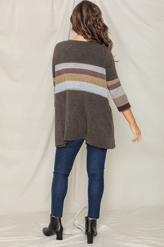 A woman with shoulder-length hair is wearing the Knit Stripe Panel A-Line Tunic, featuring horizontal brown, green, and blue knit stripe panels. She stands against a paneled white wall with one hand touching her neck and pairs the tunic with dark jeans.