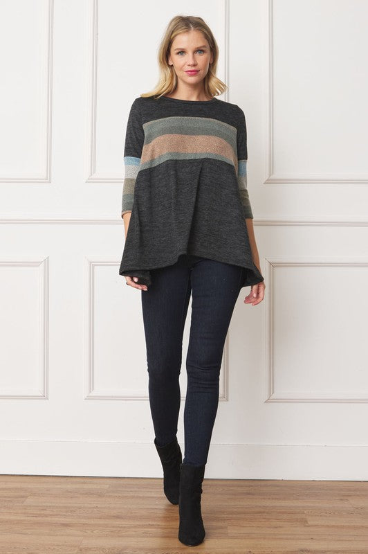 A woman with shoulder-length hair is wearing the Knit Stripe Panel A-Line Tunic, featuring horizontal brown, green, and blue knit stripe panels. She stands against a paneled white wall with one hand touching her neck and pairs the tunic with dark jeans.