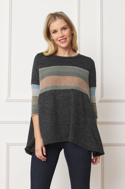 A woman with shoulder-length hair is wearing the Knit Stripe Panel A-Line Tunic, featuring horizontal brown, green, and blue knit stripe panels. She stands against a paneled white wall with one hand touching her neck and pairs the tunic with dark jeans.