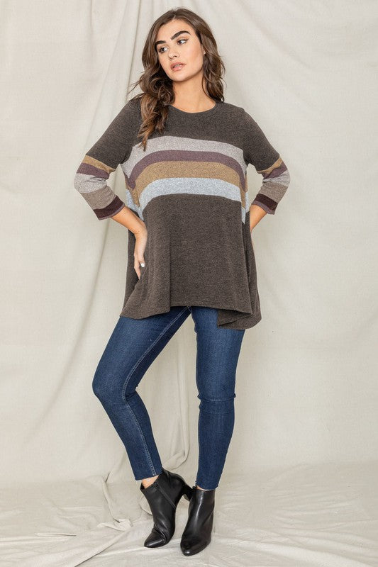 A person is standing against a beige backdrop, wearing a Plus Knit Stripe Panel A Line Tunic with horizontal stripes in various shades and blue jeans.
