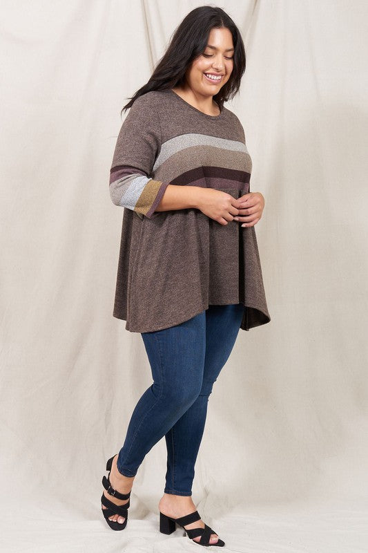 A person is standing against a beige backdrop, wearing a Plus Knit Stripe Panel A Line Tunic with horizontal stripes in various shades and blue jeans.