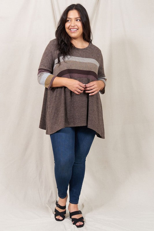 A person is standing against a beige backdrop, wearing a Plus Knit Stripe Panel A Line Tunic with horizontal stripes in various shades and blue jeans.