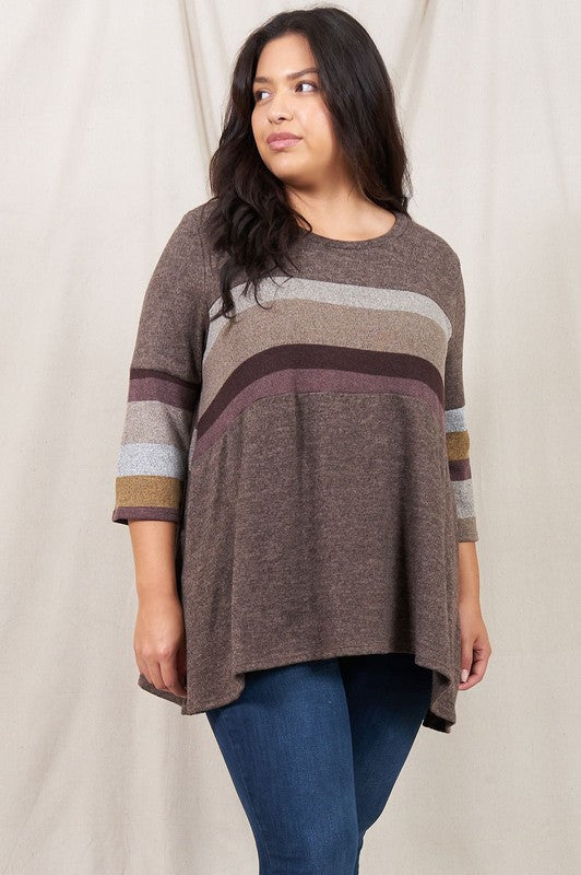 A person is standing against a beige backdrop, wearing a Plus Knit Stripe Panel A Line Tunic with horizontal stripes in various shades and blue jeans.