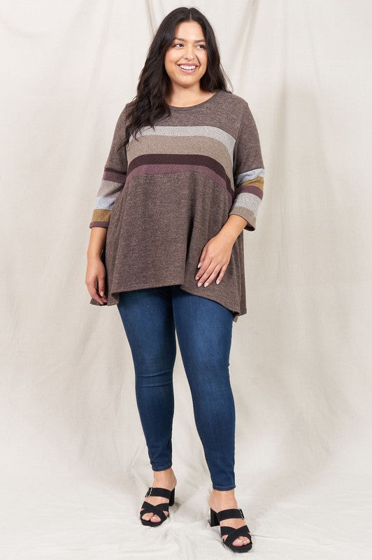 A person is standing against a beige backdrop, wearing a Plus Knit Stripe Panel A Line Tunic with horizontal stripes in various shades and blue jeans.