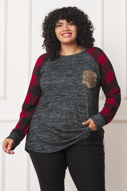 A woman with curly hair, wearing a Plus Checker Sleeve Sequin Pocket Tunic featuring dark grey fabric and red plaid checker sleeves, stands against a white background. Made in the United States, she has her hand in her pocket and is smiling.