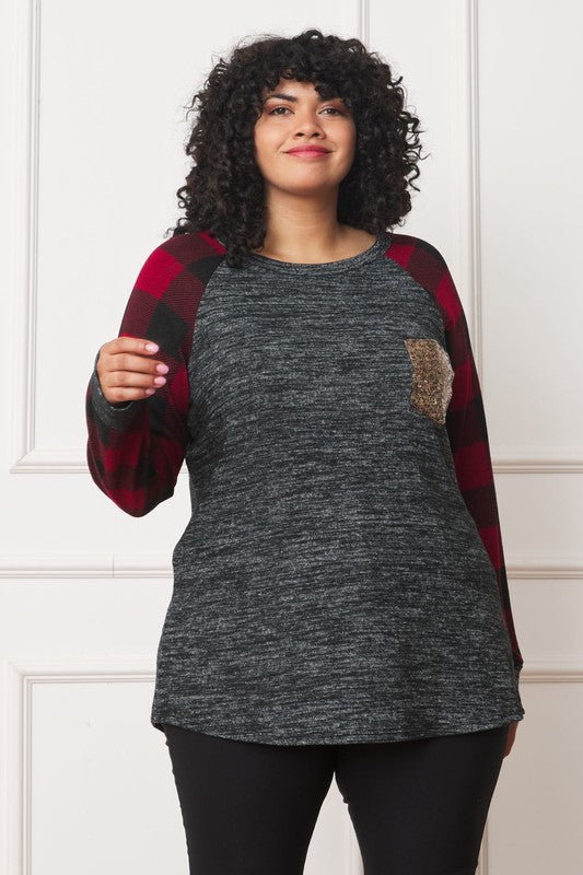 A woman with curly hair, wearing a Plus Checker Sleeve Sequin Pocket Tunic featuring dark grey fabric and red plaid checker sleeves, stands against a white background. Made in the United States, she has her hand in her pocket and is smiling.