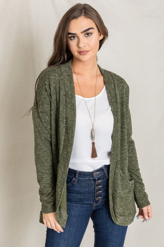 A woman with long dark hair is wearing a green Solid Drape Pocket Cardigan, along with a white shirt and blue jeans, posing against a plain background.