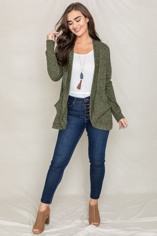 A woman with long dark hair is wearing a green Solid Drape Pocket Cardigan, along with a white shirt and blue jeans, posing against a plain background.