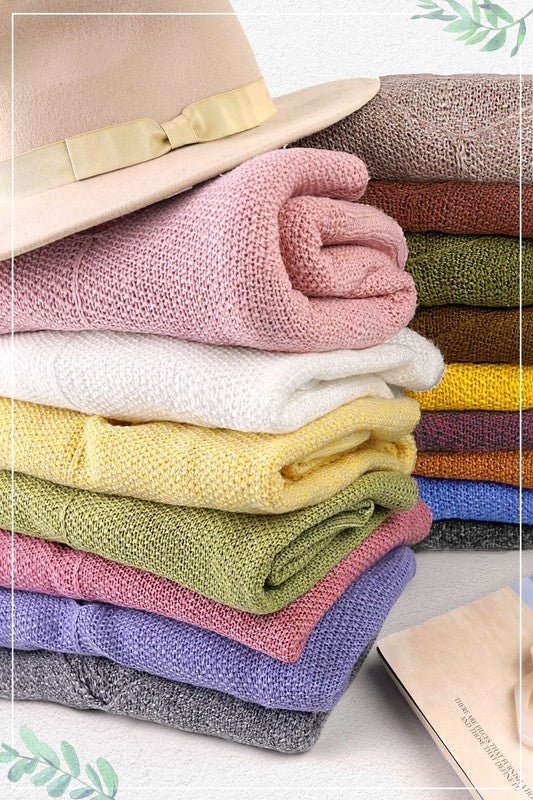 A person wearing a red beret, the gray Winnie Sweater in a relaxed off-shoulder fit, distressed denim shorts, and gold jewelry holds a plaid cloth while posing against a beige background.