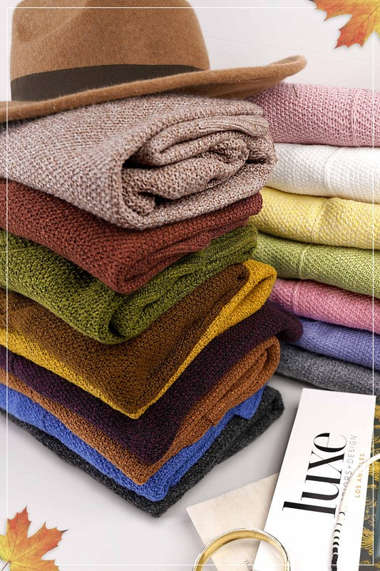 A person wearing a red beret, the gray Winnie Sweater in a relaxed off-shoulder fit, distressed denim shorts, and gold jewelry holds a plaid cloth while posing against a beige background.