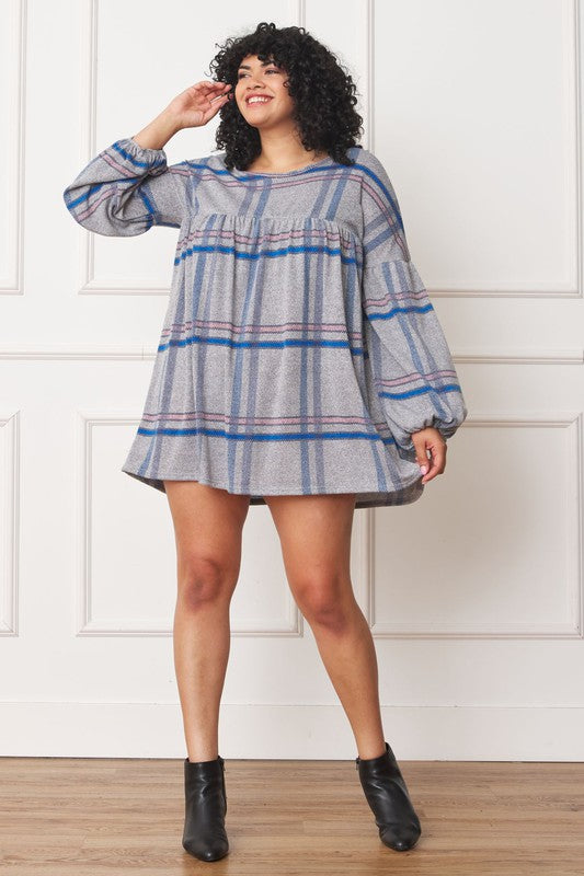 A person with curly hair wearing a Plus Plaid Empire Waist Balloon Sleeve Mini Dress stands against a white wall, smiling.