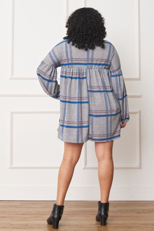 A person with curly hair wearing a Plus Plaid Empire Waist Balloon Sleeve Mini Dress stands against a white wall, smiling.
